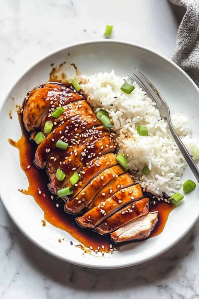Tender baked teriyaki chicken slices coated in glossy sauce, sprinkled with sesame seeds and chopped green onions, served with white rice on a round plate.