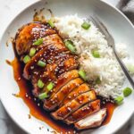 Tender baked teriyaki chicken slices coated in glossy sauce, sprinkled with sesame seeds and chopped green onions, served with white rice on a round plate.