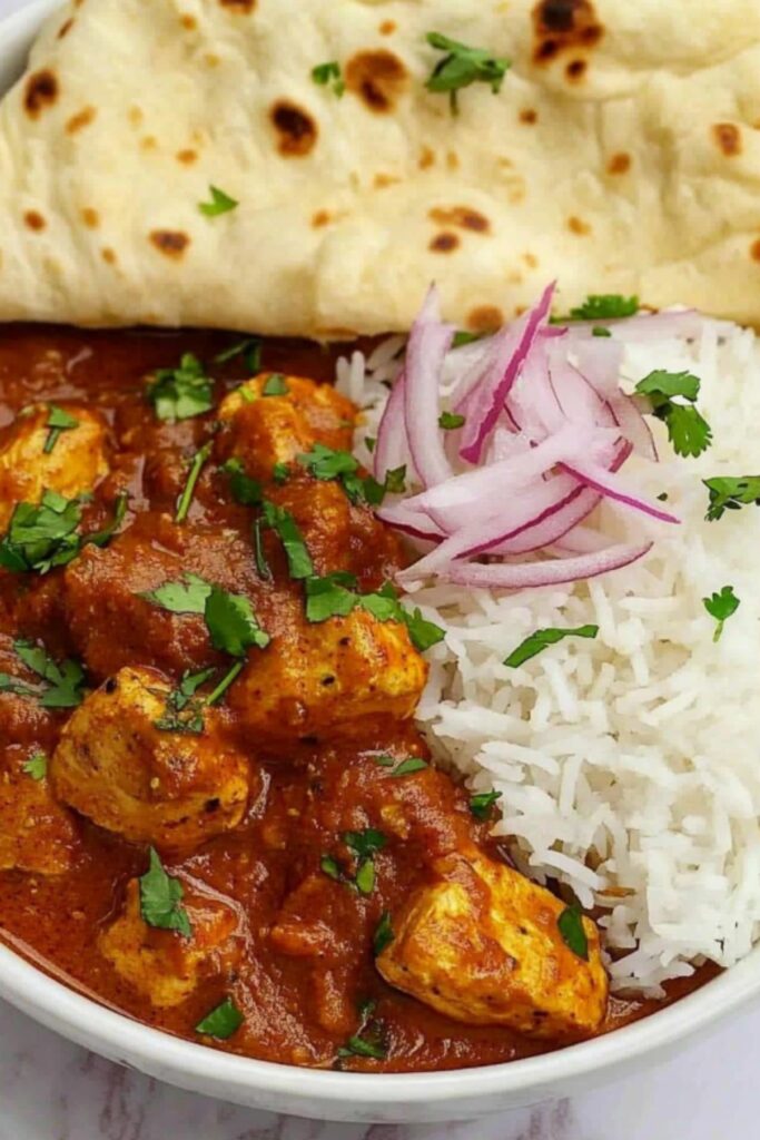 Delicious boneless chicken curry served with white basmati rice, naan bread, and fresh red onion slices, garnished with cilantro.