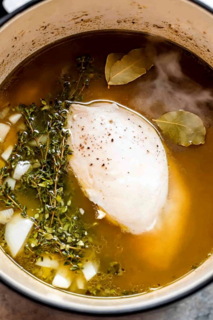 A pot with simmering chicken breast, bay leaves, fresh thyme, and diced onions in a rich chicken broth.