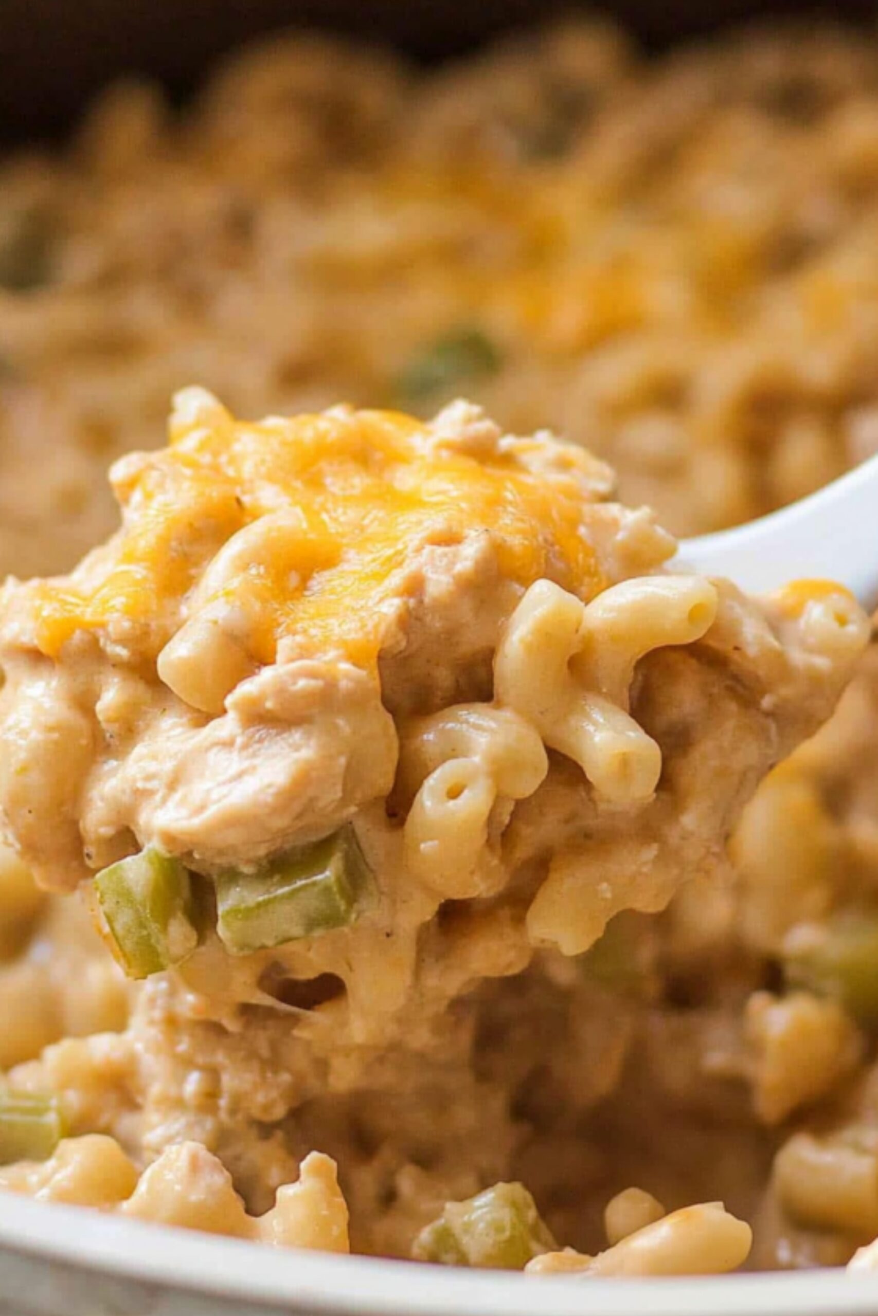 A white spoon lifting a cheesy, creamy bite of chicken macaroni casserole with green bell peppers.