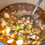 A ladle scooping hearty high protein chicken noodle soup with tender chicken, beans, celery, and flavorful broth