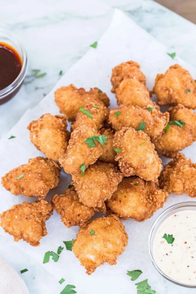 Freshly fried crispy chicken nuggets sprinkled with parsley, served on parchment paper with ranch and barbecue sauce bowls