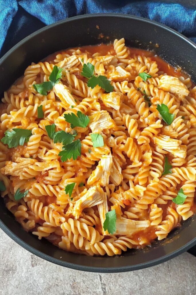 A black skillet filled with fusilli pasta, shredded leftover roasted chicken, and a rich tomato sauce, garnished with fresh parsley.