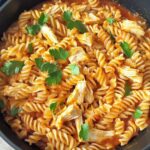 A black skillet filled with fusilli pasta, shredded leftover roasted chicken, and a rich tomato sauce, garnished with fresh parsley.