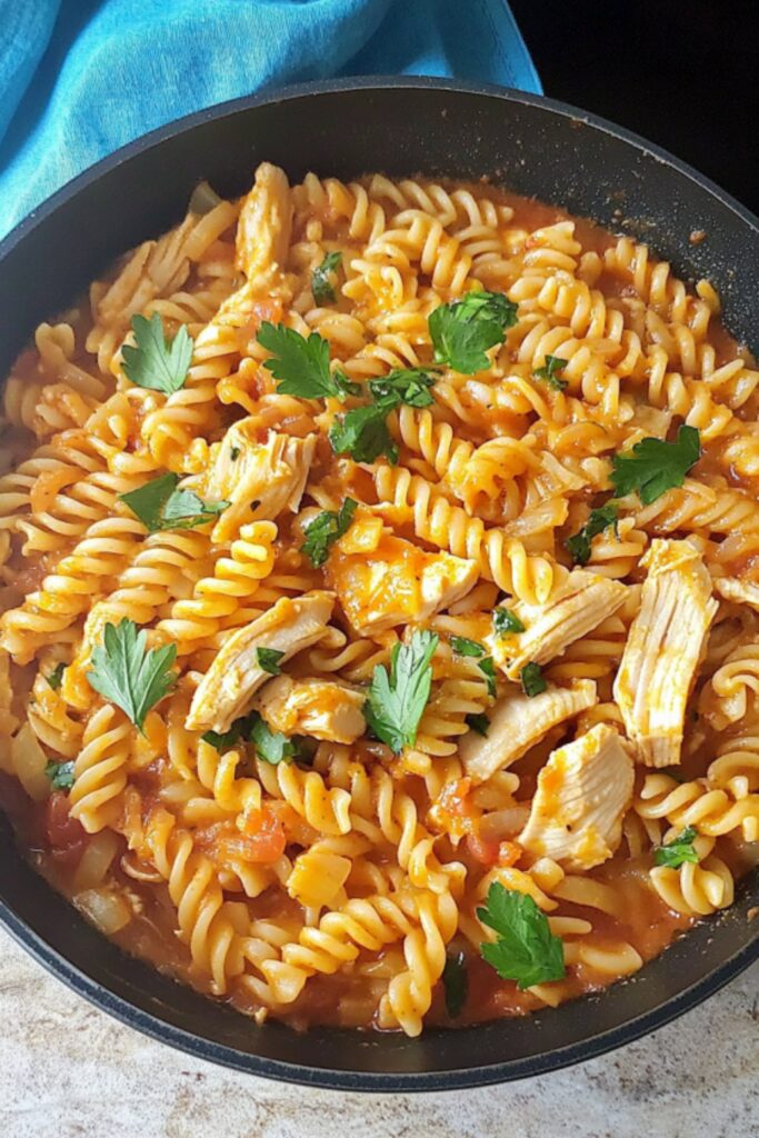 A close-up of fusilli pasta with shredded chicken in a tomato-based sauce, garnished with fresh parsley, served in a black skillet.