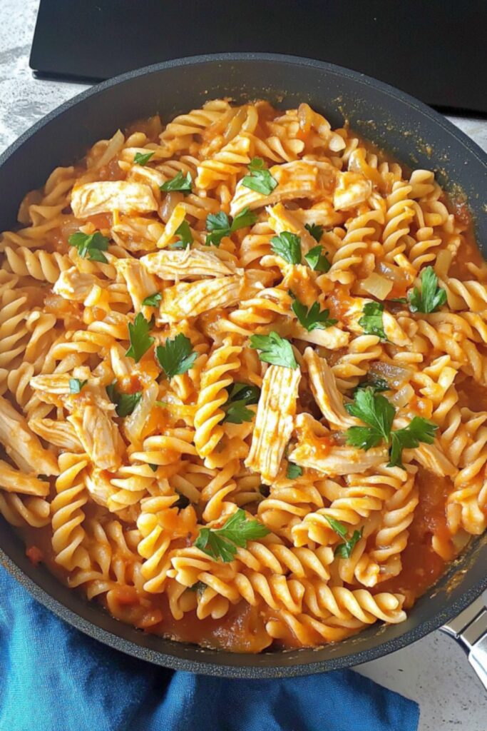 A skillet of fusilli pasta mixed with shredded leftover roasted chicken in a rich tomato sauce, topped with fresh parsley for garnish.