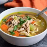 A close-up top-down view of a white bowl filled with low FODMAP chicken soup, featuring shredded chicken, carrots, celery, and mushrooms, garnished with fresh parsley.