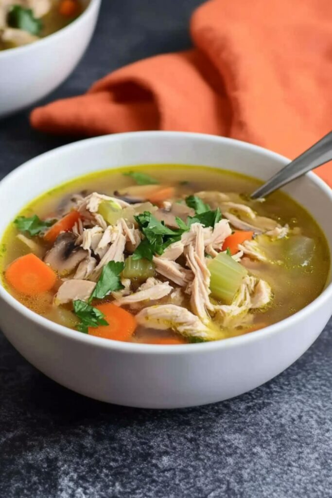 A close-up top-down view of a white bowl filled with low FODMAP chicken soup, featuring shredded chicken, carrots, celery, and mushrooms, garnished with fresh parsley.
