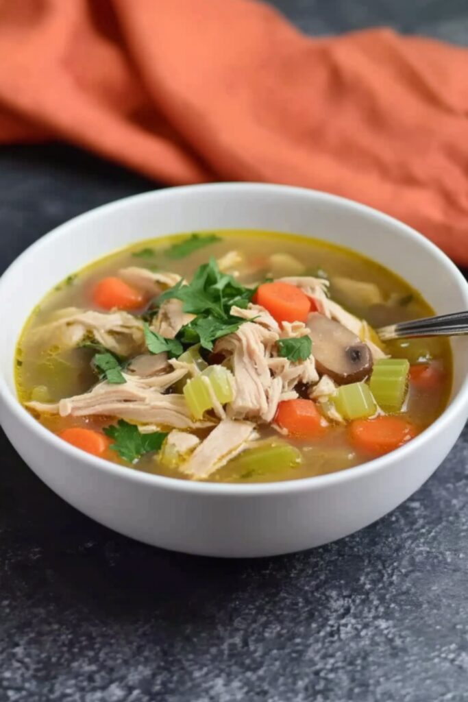 A close-up side view of a bowl of low FODMAP chicken soup, showing shredded chicken, carrots, celery, and mushrooms in a rich golden broth with a spoon resting inside.