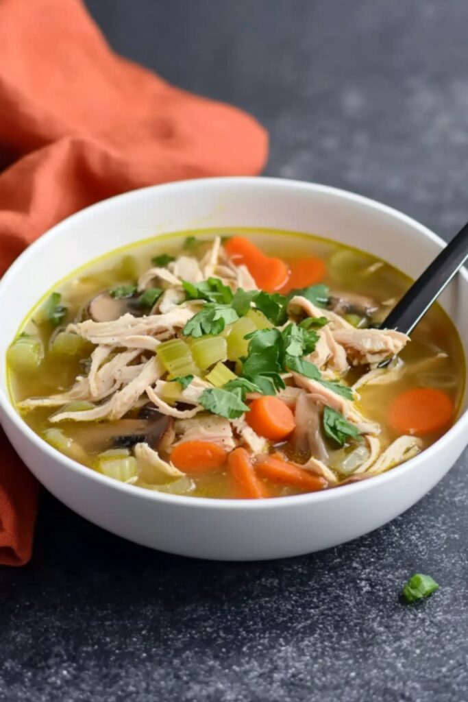 A warm bowl of low FODMAP chicken soup with shredded chicken, mushrooms, carrots, and celery, served on a dark countertop with an orange cloth in the background.