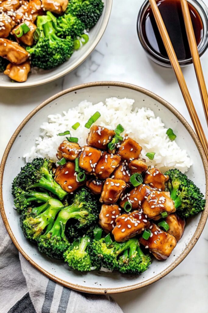 Overhead view of a teriyaki chicken bowl featuring white rice, tender chicken pieces, steamed broccoli, sesame seeds, and fresh green onions.