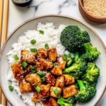 Bowl of fluffy white rice topped with leftover teriyaki chicken pieces, steamed broccoli, and garnished with sesame seeds and green onions.