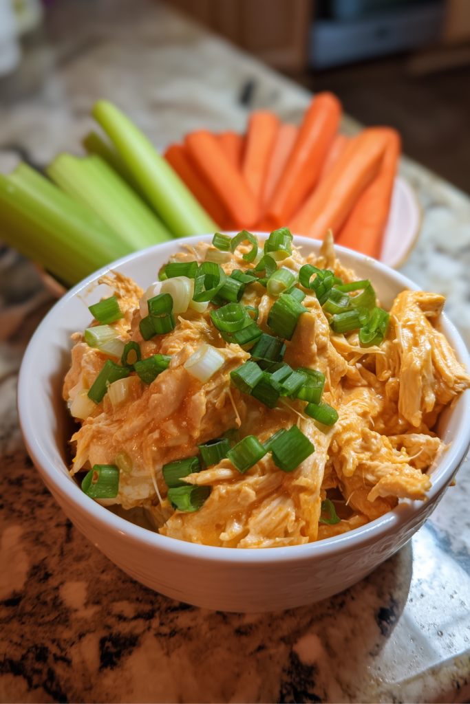 Bowl of shredded low sodium buffalo chicken topped with green onions, served with celery and carrot sticks on a marble countertop