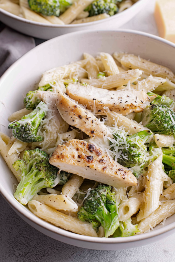 Close-up of grilled chicken slices over creamy Alfredo penne pasta with broccoli in a white bowl