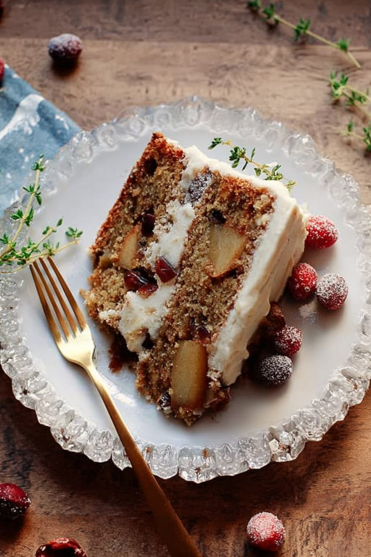 Slice of frosted Thanksgiving layer cake with apples, cranberries, and pecans, garnished with sugared cranberries and thyme sprigs.