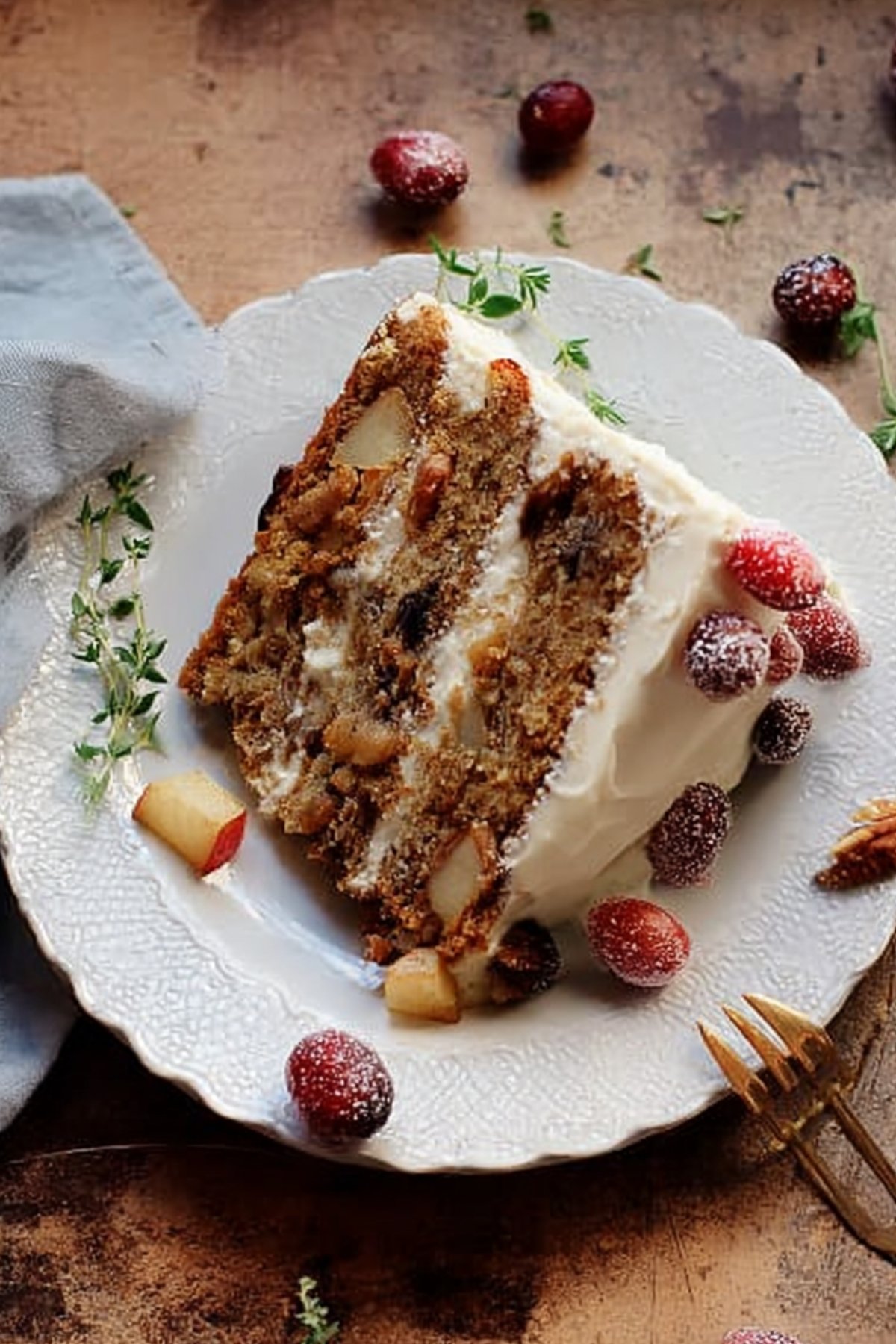 Close-up of moist Thanksgiving layer cake slice with frosting, apples, cranberries, and pecans on a plate with herbs.
