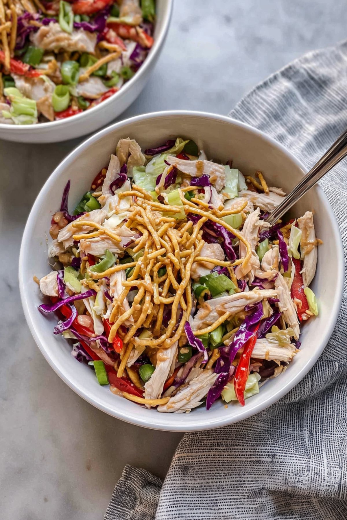 Bowl of Chinese chicken salad topped with crunchy chow mein noodles beside a striped napkin.