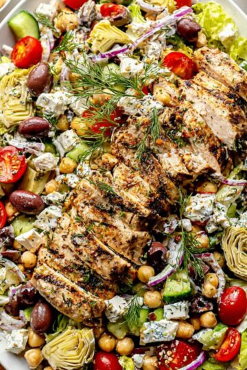 Overhead photo of a large platter of Greek chicken salad with sliced chicken breast, fresh vegetables, and creamy feta dressing.