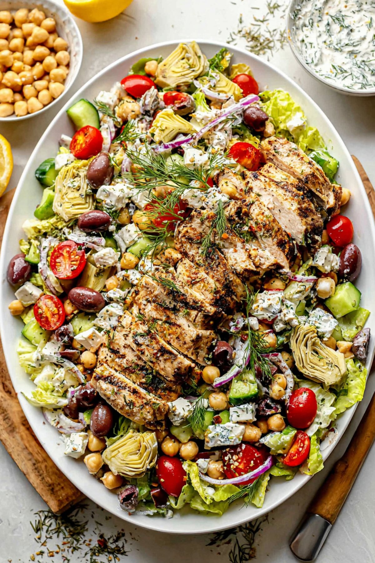 Greek chicken salad served on a platter with sliced chicken breast, fresh dill, artichokes, cucumbers, tomatoes, and creamy feta dressing.
