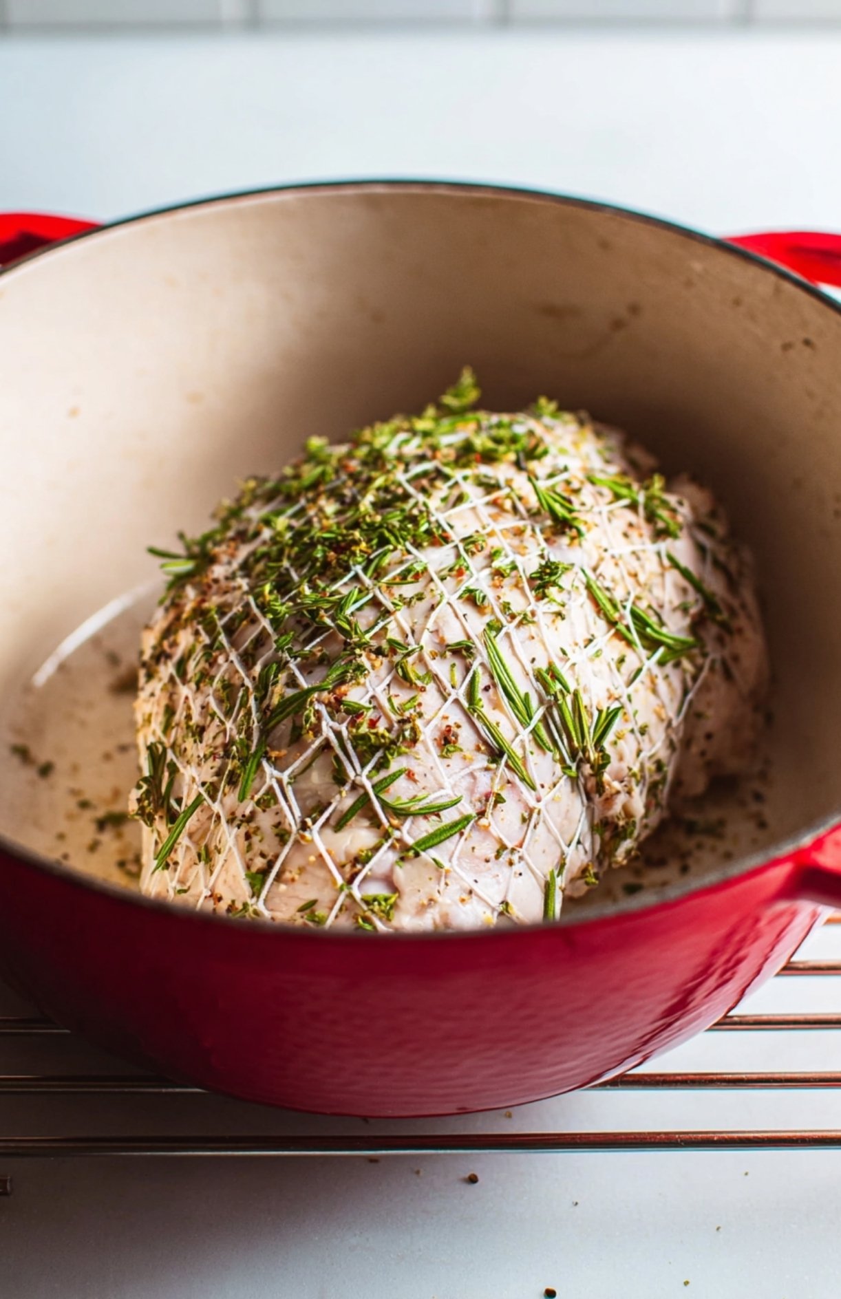 Uncooked boneless Butterball turkey roast covered in rosemary inside a red Dutch oven baking in the oven.