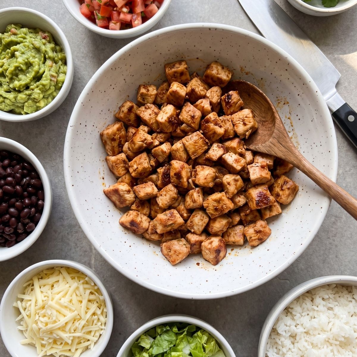 A bowl of diced chipotle seasoned chicken surrounded by meal prep sides like guacamole, black beans, and white rice.