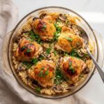 Baked chicken and rice casserole featuring five golden brown chicken thighs over fluffy rice and sliced mushrooms in a round glass dish, garnished with fresh parsley.