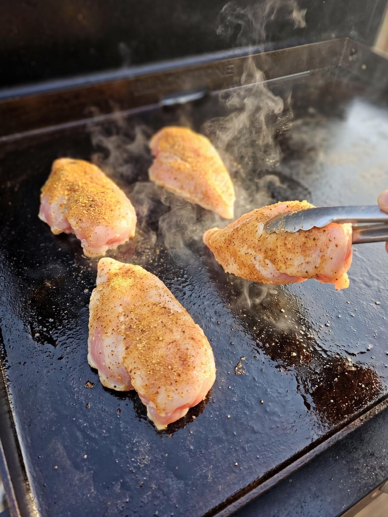 Seasoned chicken breasts searing on a hot Blackstone griddle with visible steam rising from the flat top.