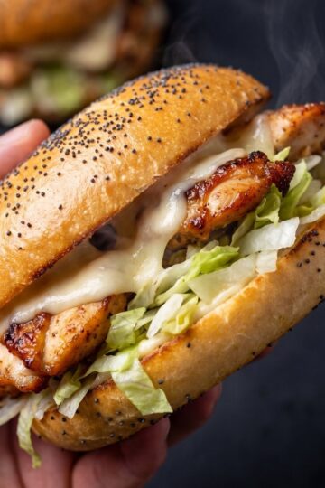 A hand holding a toasted poppy seed bun filled with grilled chicken, melted cheese, and shredded lettuce.