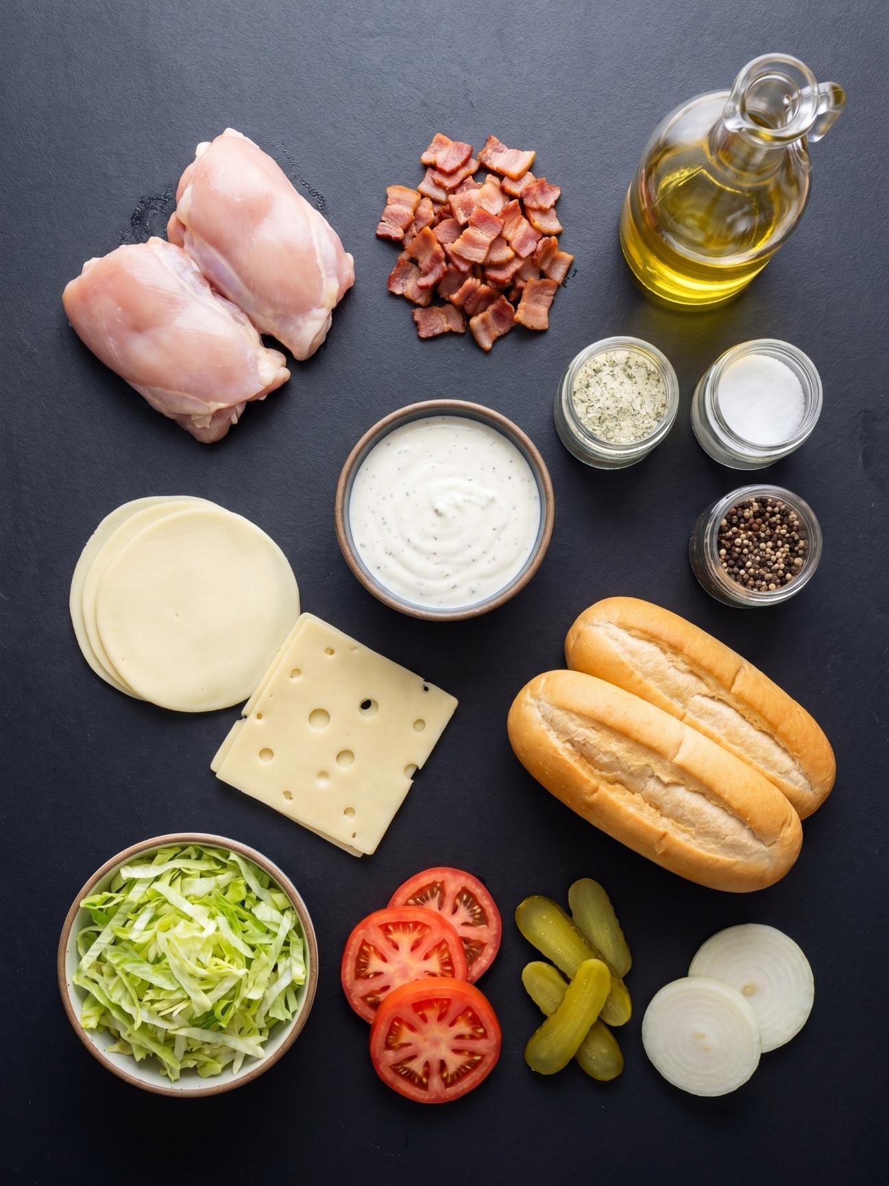 Top down view of ingredients including raw chicken, chopped bacon, ranch dressing, cheese slices, rolls, and fresh vegetables on a dark surface.