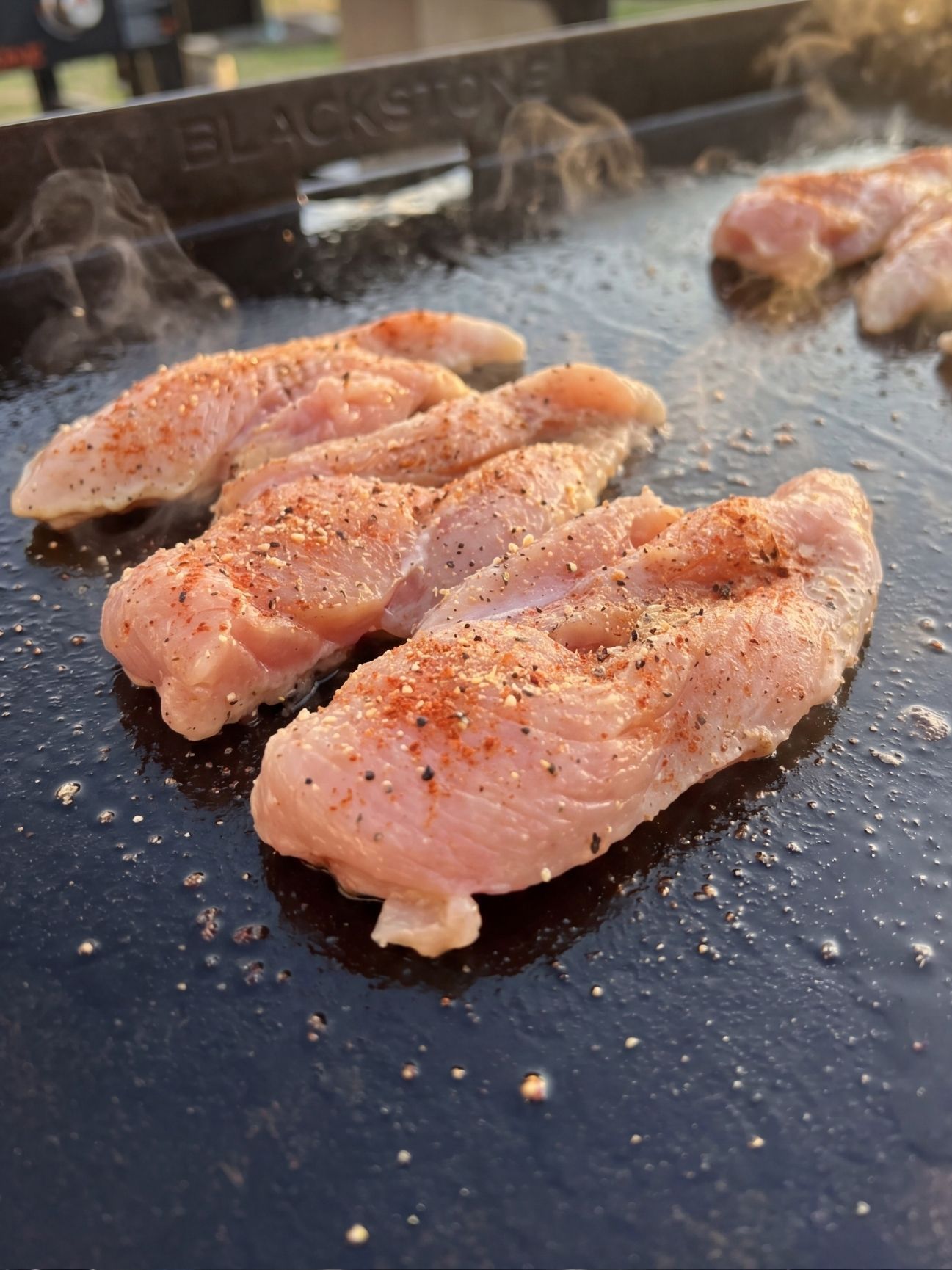 Sliced raw chicken thighs seasoned with red and black spices cooking on a hot oiled griddle.