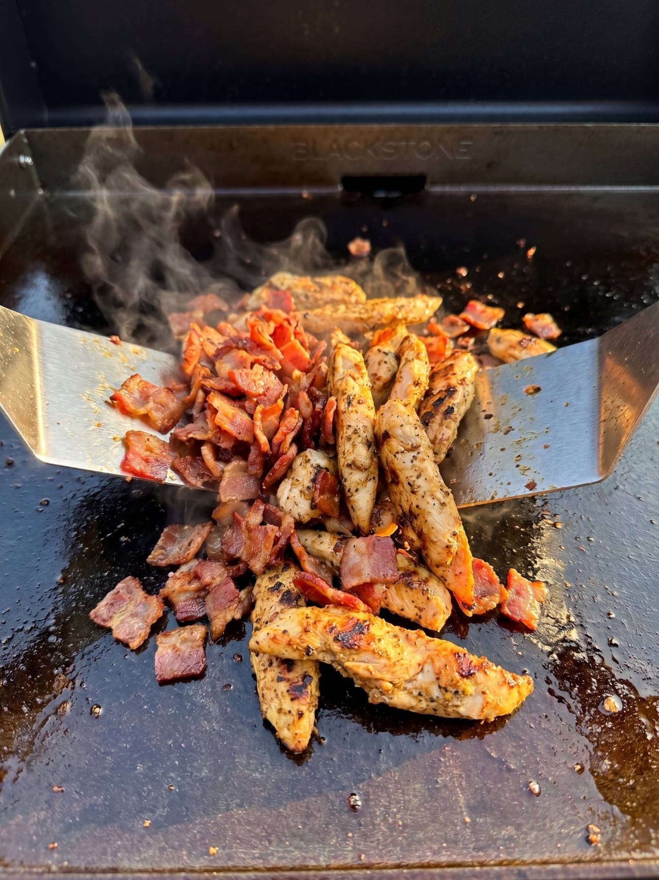 Two metal spatulas mixing together grilled chicken strips and crispy chopped bacon on a Blackstone griddle.