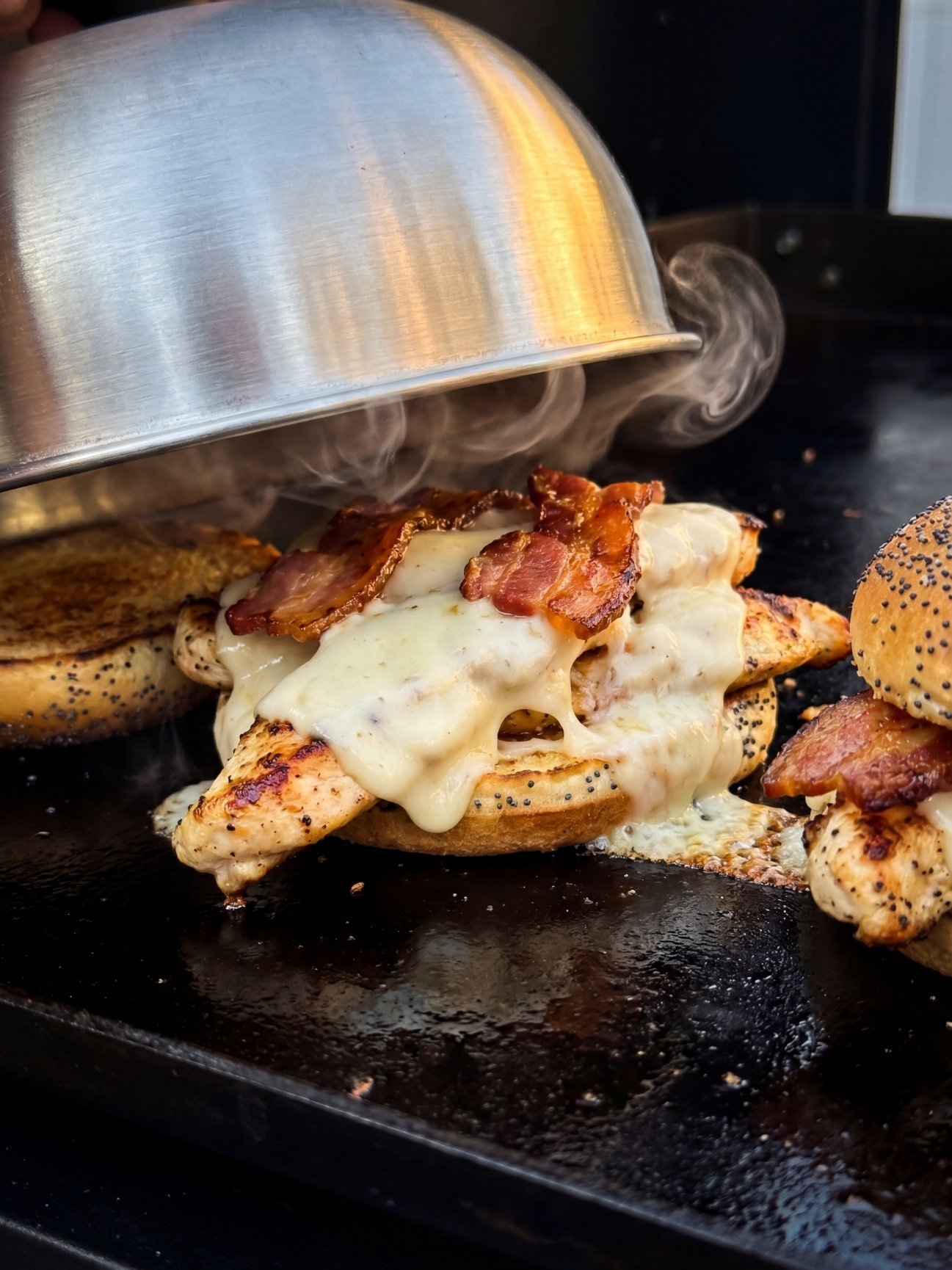 A metal melting dome being lifted to reveal a sandwich with melted white cheese and bacon on the griddle.