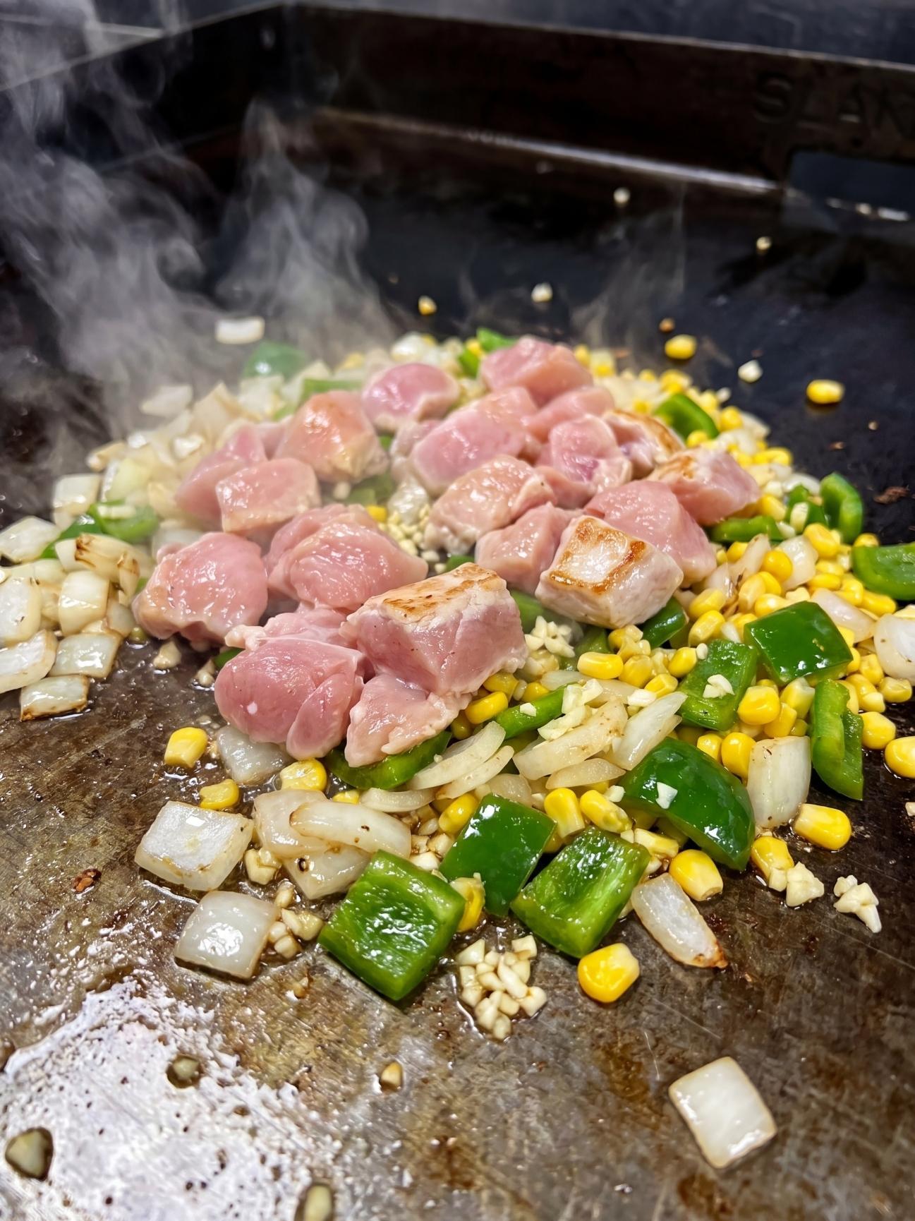 Raw diced chicken cooking on a griddle next to green peppers, onions, and yellow corn.