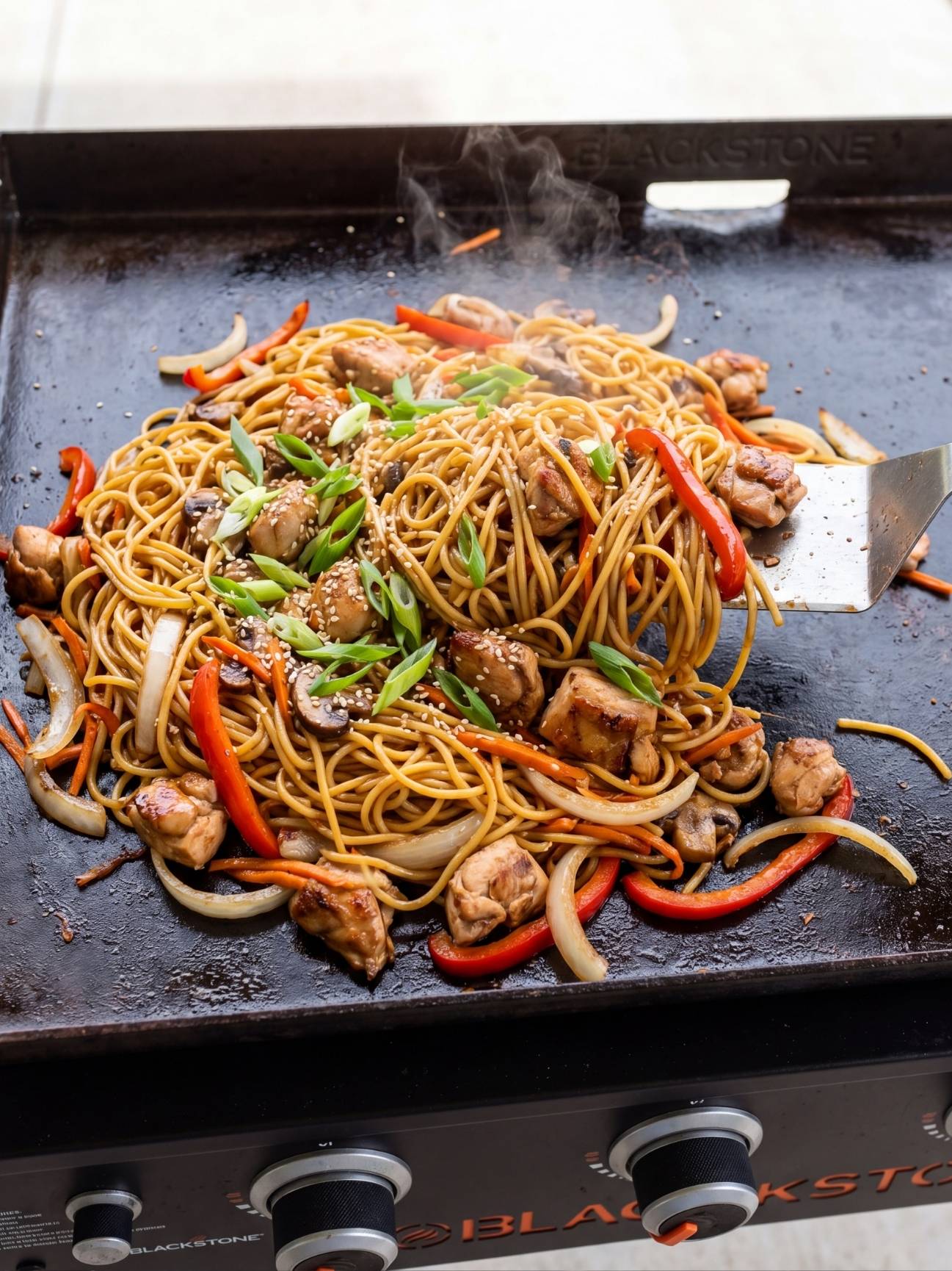 Tossed and finished Blackstone chicken lo mein, ready to serve, garnished with green onions and sesame seeds.