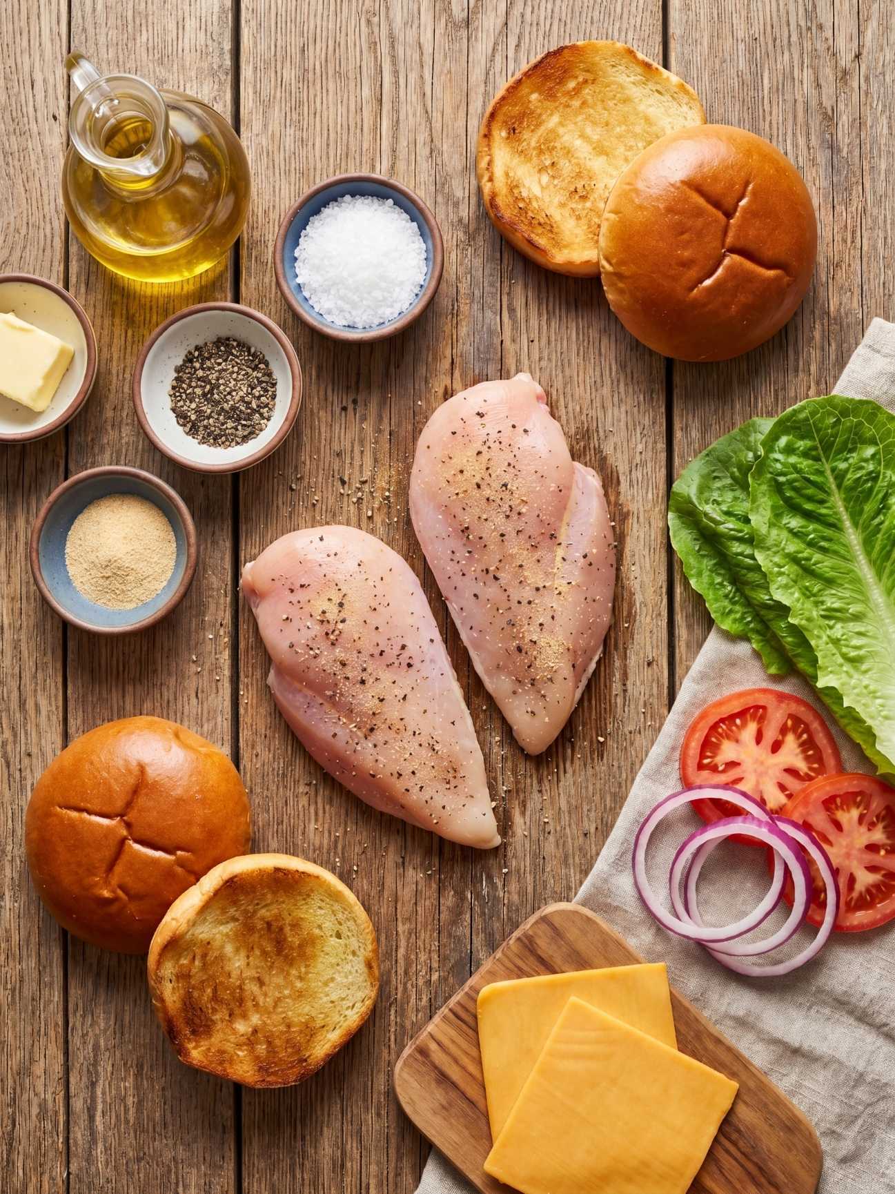 Overhead view of raw seasoned chicken breasts, brioche buns, sliced cheese, lettuce, tomato, and red onion on a wooden board.