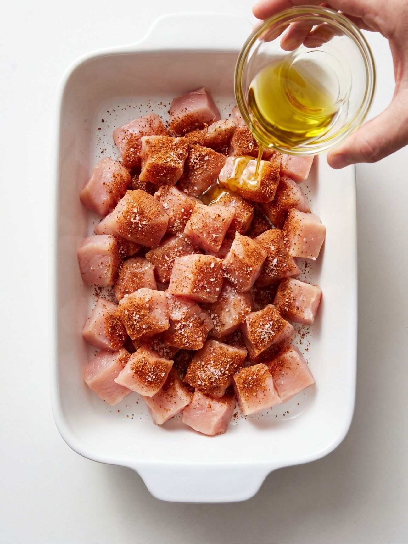 Diced raw chicken in a white baking dish being drizzled with avocado oil and taco seasoning.