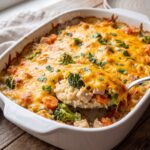 A white baking dish filled with a cheesy chicken and rice bake with vegetables, showing broccoli and carrots under a melted cheddar cheese crust.