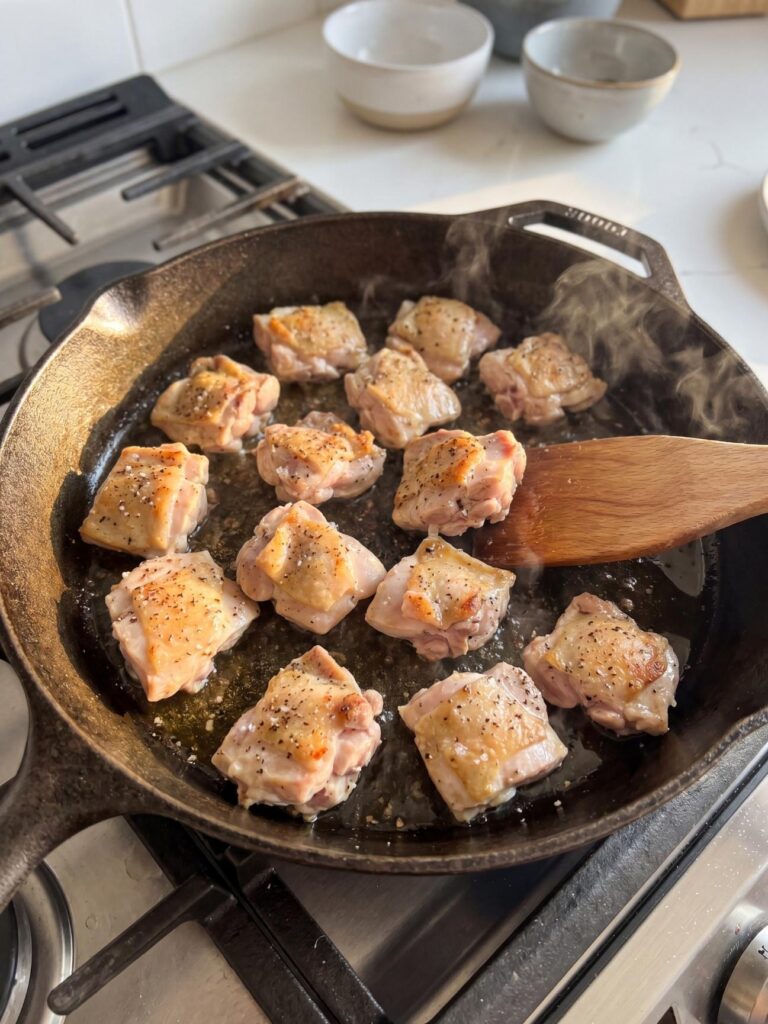 Cubed chicken breast pieces being seared in a hot cast iron skillet with a wooden spatula turning them over.