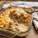 A glass baking dish filled with a creamy chicken and rice casserole, with a large spoonful showing tender chicken and melted cheese being lifted from the pan.