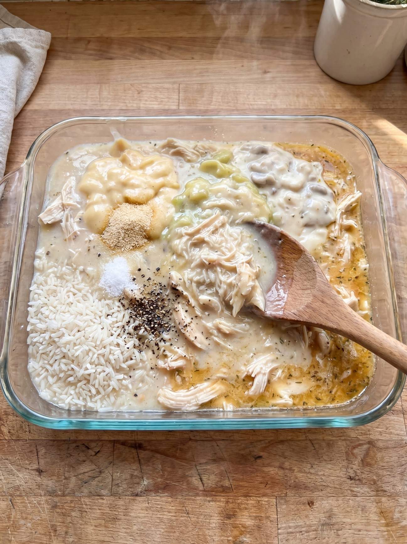 A wooden spoon mixing raw white rice, shredded chicken, cream soups, and spices together inside a clear glass baking dish.