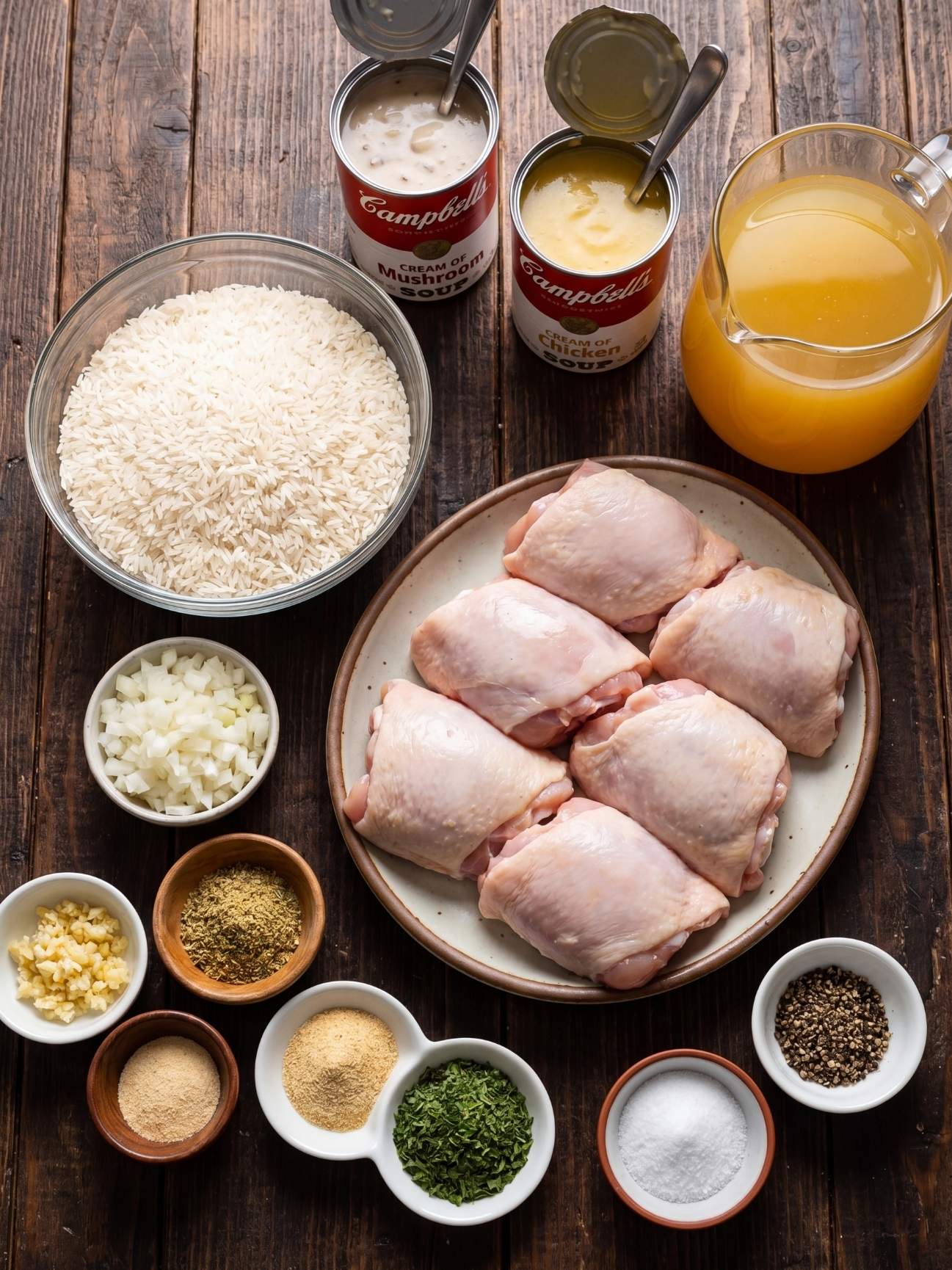 Overhead view of raw ingredients for chicken and rice bake including chicken thighs, long grain white rice, cream of mushroom soup, cream of chicken soup, chicken broth, and individual bowls of spices on a dark wooden background
