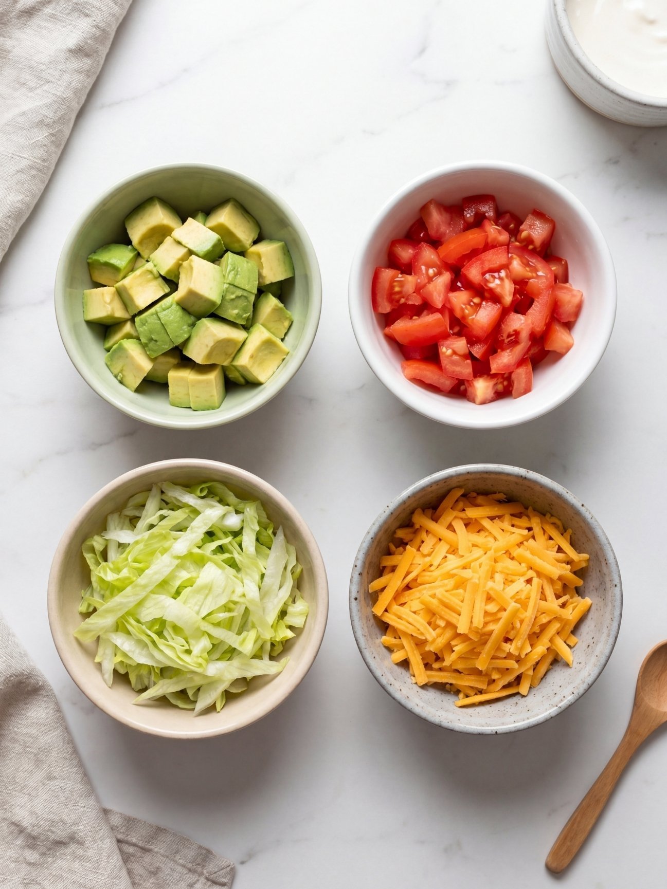 Four small white bowls containing diced avocado, chopped tomatoes, shredded lettuce, and grated cheddar cheese on a white marble surface.
