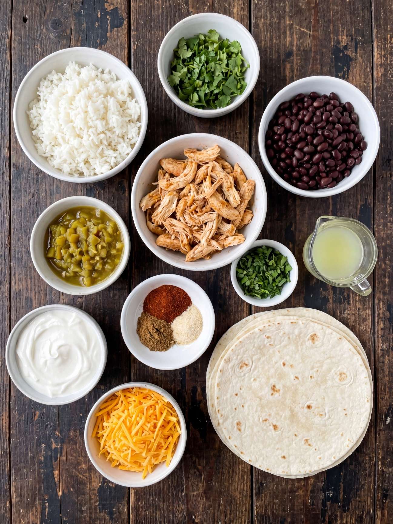 Overhead view of white bowls filled with southwest black bean chicken wrap ingredients including shredded chicken, black beans, cooked rice, shredded cheese, and green chilies on a dark wooden surface