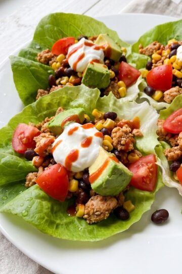 Four Southwest chicken lettuce wraps served on a white plate topped with diced avocado, fresh tomatoes, sour cream, and hot sauce