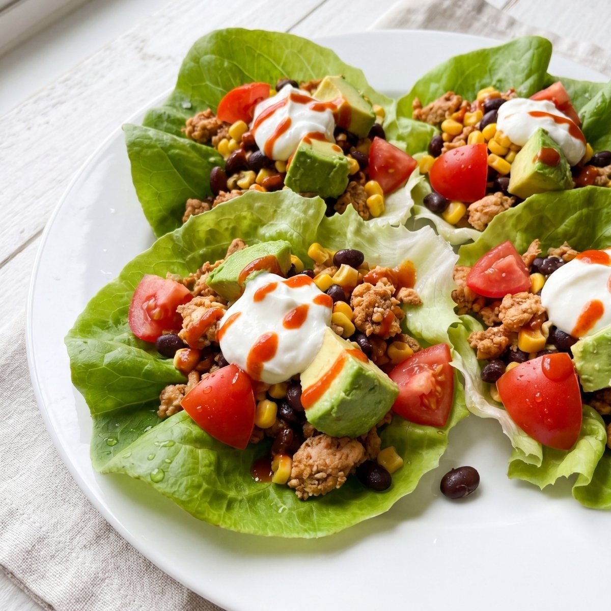 Four Southwest chicken lettuce wraps served on a white plate topped with diced avocado, fresh tomatoes, sour cream, and hot sauce